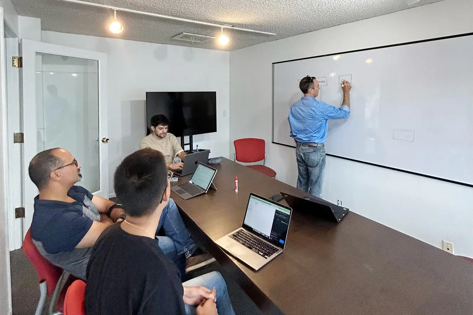 In a problem-solving meeting, one employee writes on a whiteboard while three others sit with laptops, all attentive to the presenter.