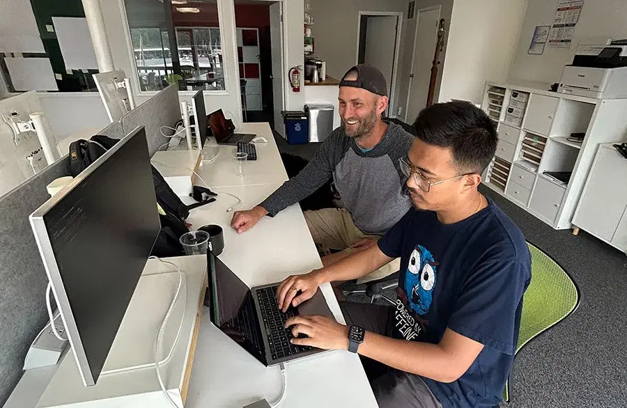 In workspace, One employee works on a laptop while another guides, creating innovative solutions collaboratively.