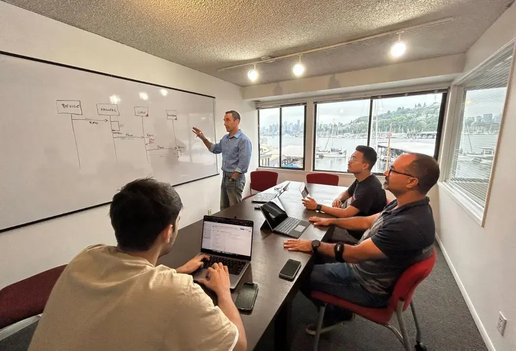 Team meeting in a conference room, three employees seated at a table, one writing on a whiteboard.