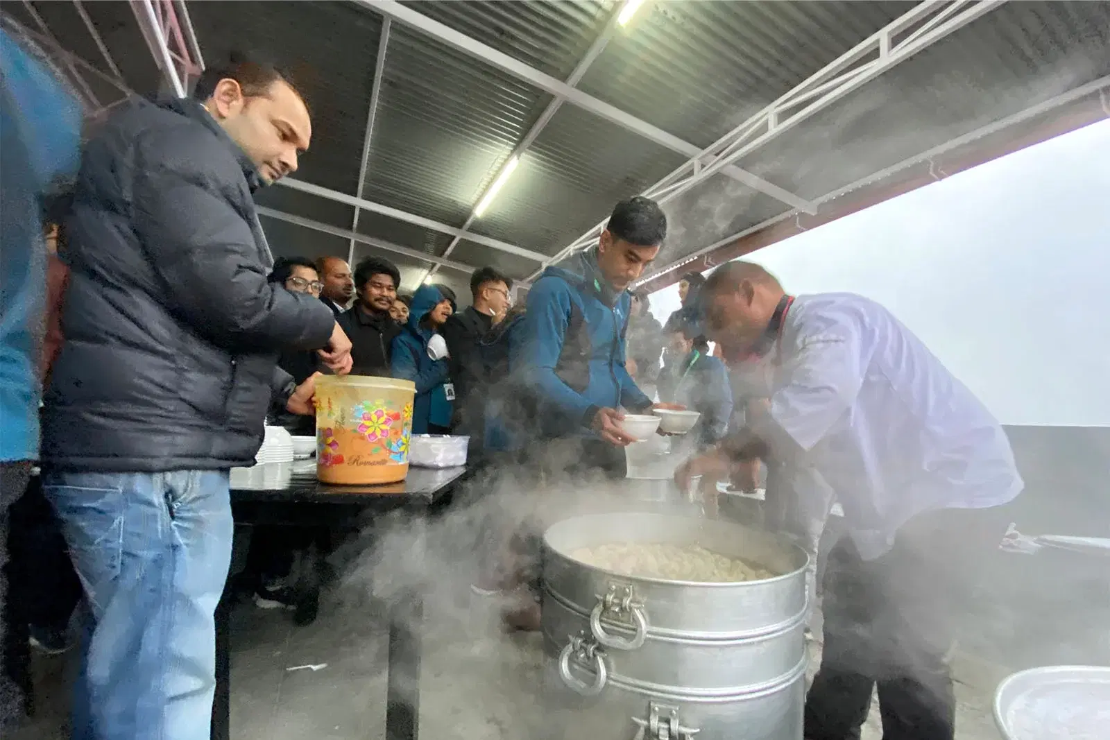 A person serving hot momos from steamers with employees lined up to enjoy the delicious treat.