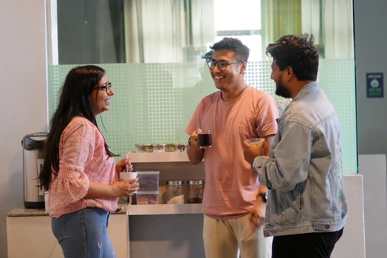 Female and two male employees enjoying coffee at a station, smiling as they socialize.