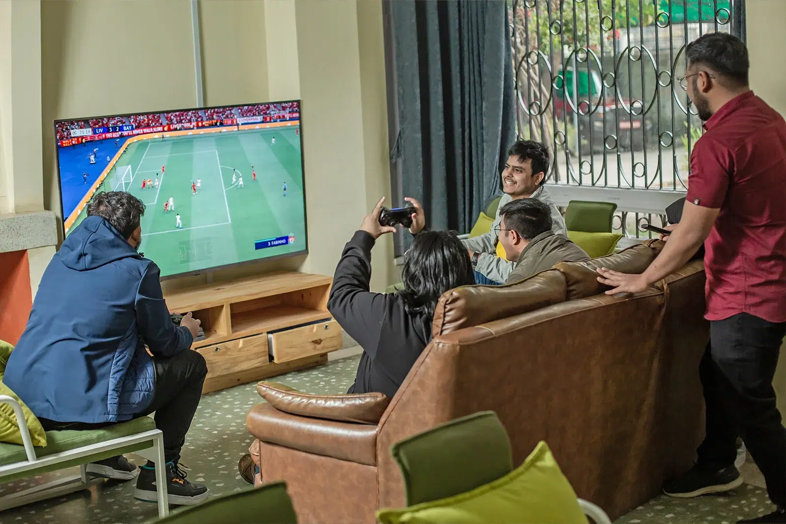Five employees playing FIFA on a large office screen: one standing, four seated on a couch.